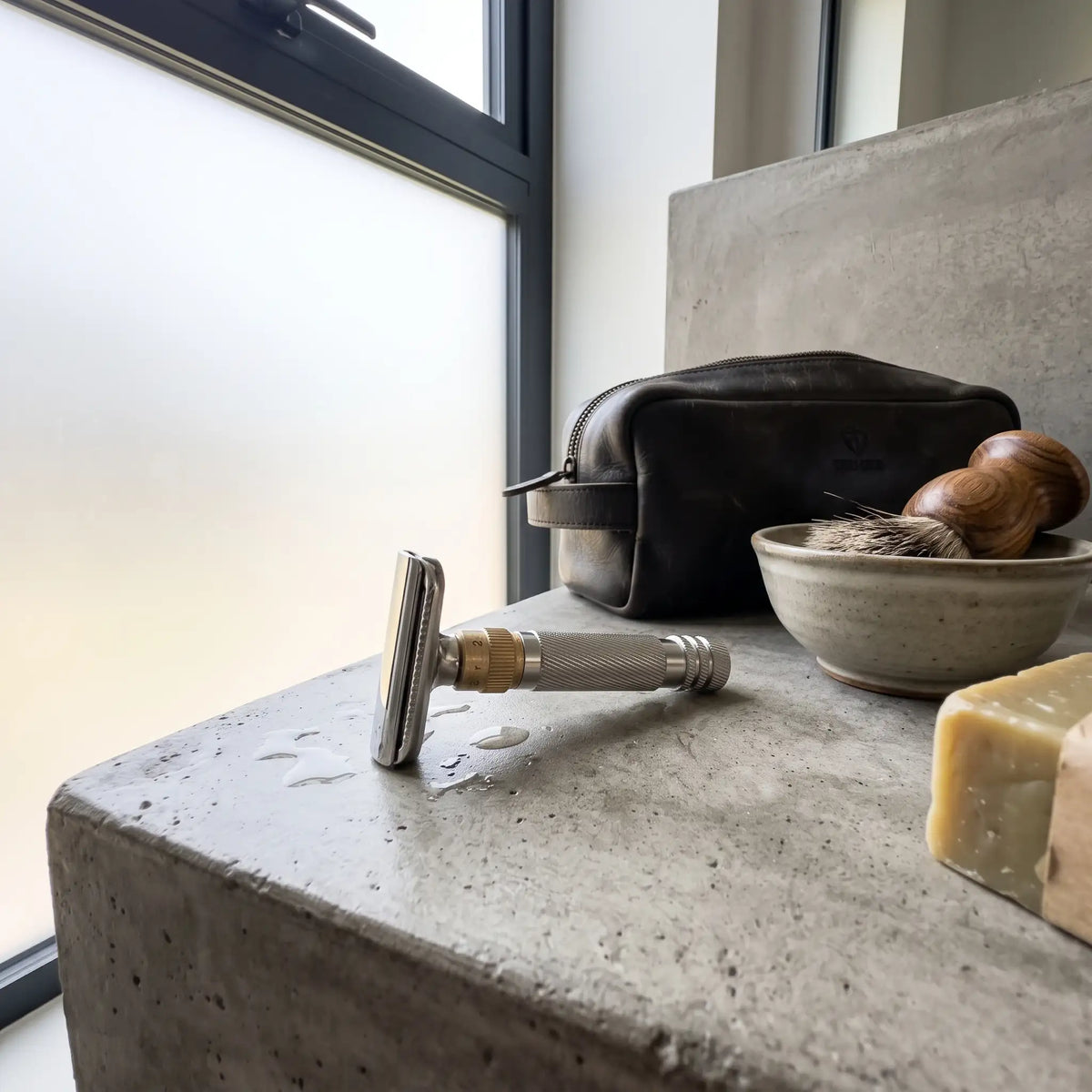 LIFER Open-Comb safety razor with brass adjustment dial resting on a concrete surface beside a leather dopp kit, wooden shaving brush, and bar of soap