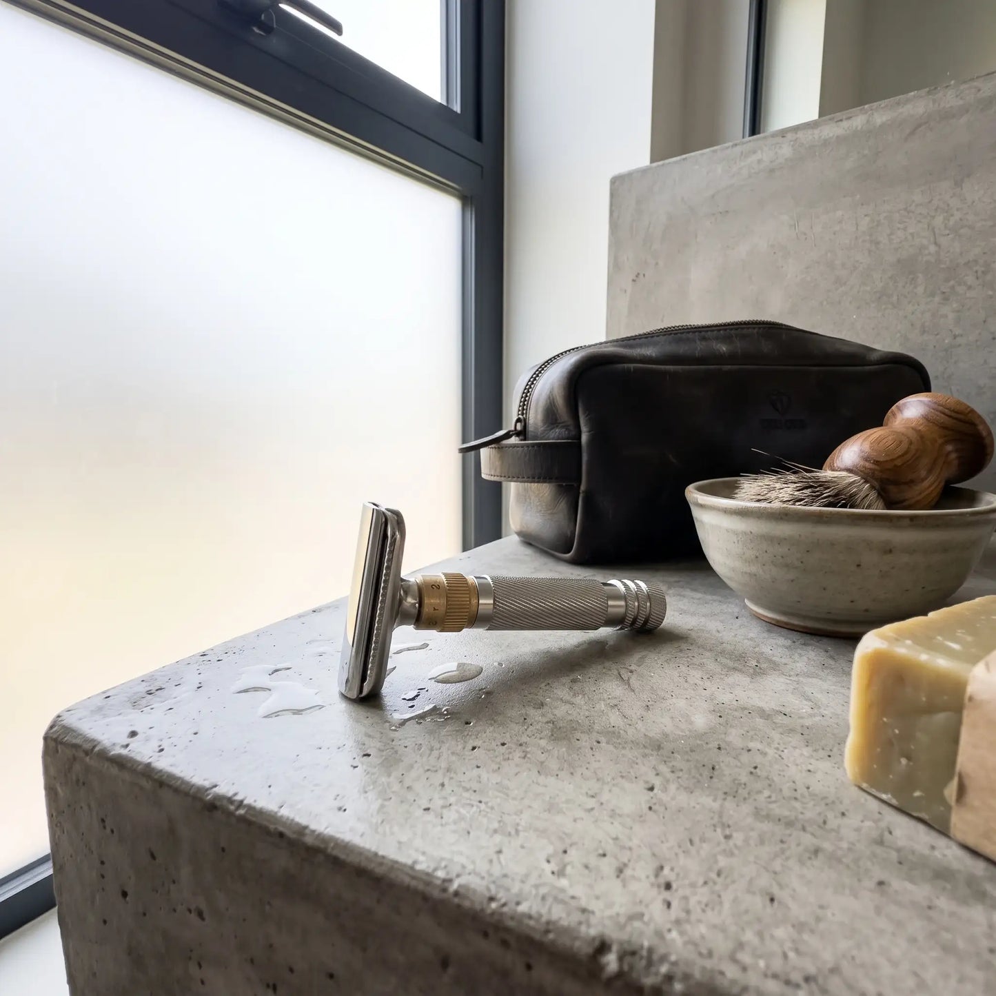 LIFER Open-Comb safety razor with brass adjustment dial resting on a concrete surface beside a leather dopp kit, wooden shaving brush, and bar of soap